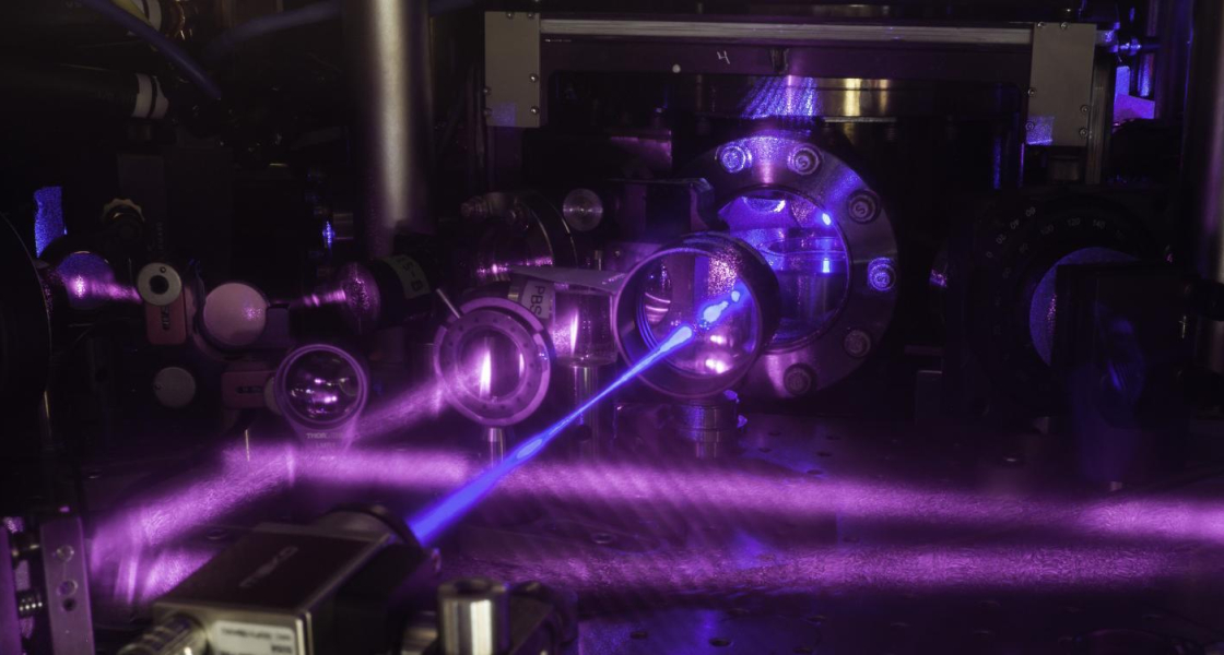 JILA's three-dimensional (3-D) quantum gas atomic clock consists of a grid of light formed by three pairs of laser beams. Multiple lasers of various colors are used to cool the atoms, trap them in a grid of light, and probe them for clock operation. A blue laser beam excites a cube-shaped cloud of strontium atoms. Strontium atoms fluorescence strongly when excited with blue light, as seen in the upper right corner behind the vacuum window.